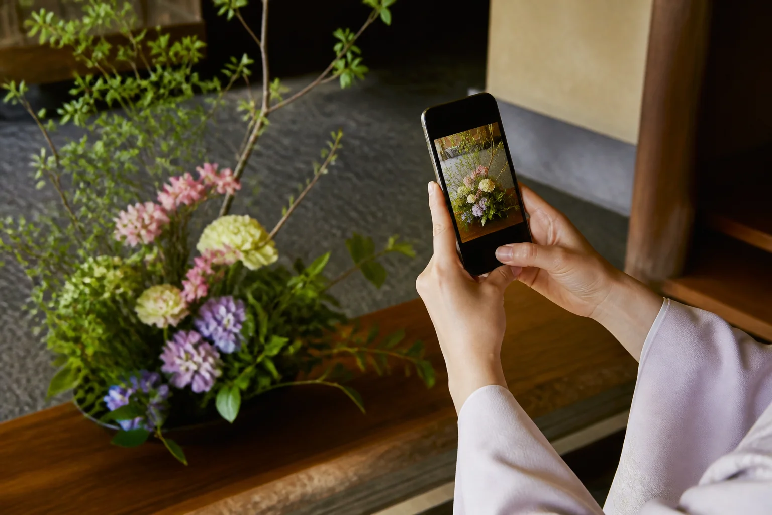 旅館の玄関に生けた季節の花を、スマートフォンで撮影している女将の手元をイメージした写真