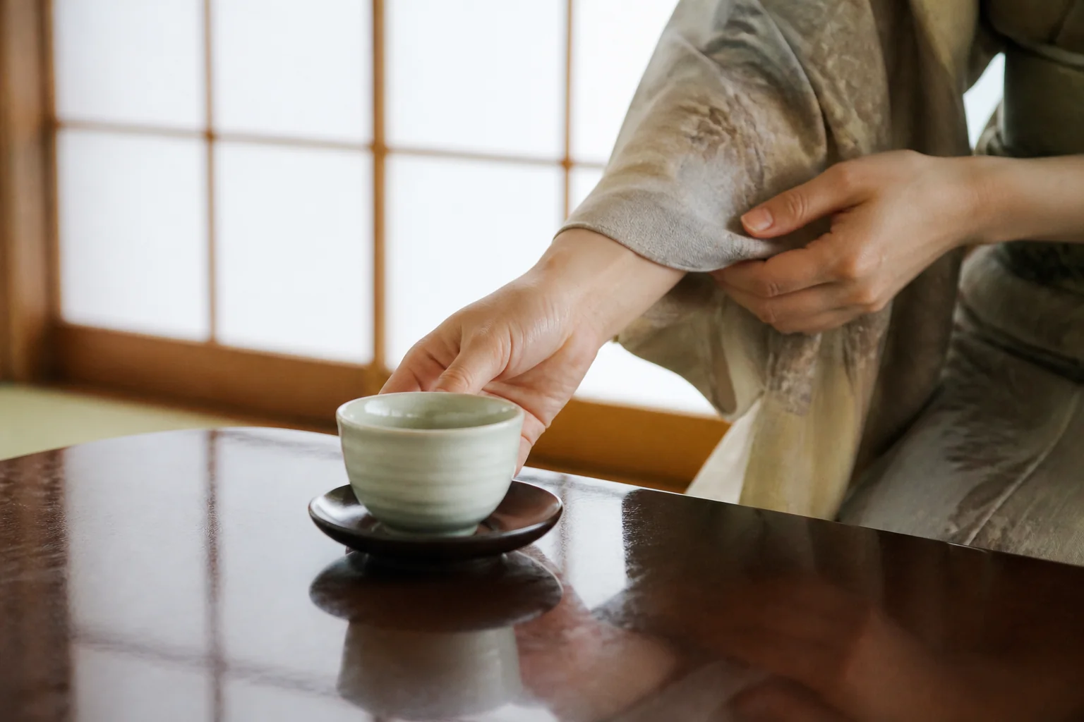 旅館の客室で、着物の袖口を押さえながら座卓に湯呑みを置く手元をイメージした写真